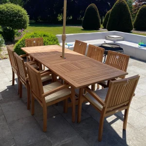 Outdoor wooden dining table with eight chairs on a stone patio, and a central umbrella, in a sunny backyard setting.